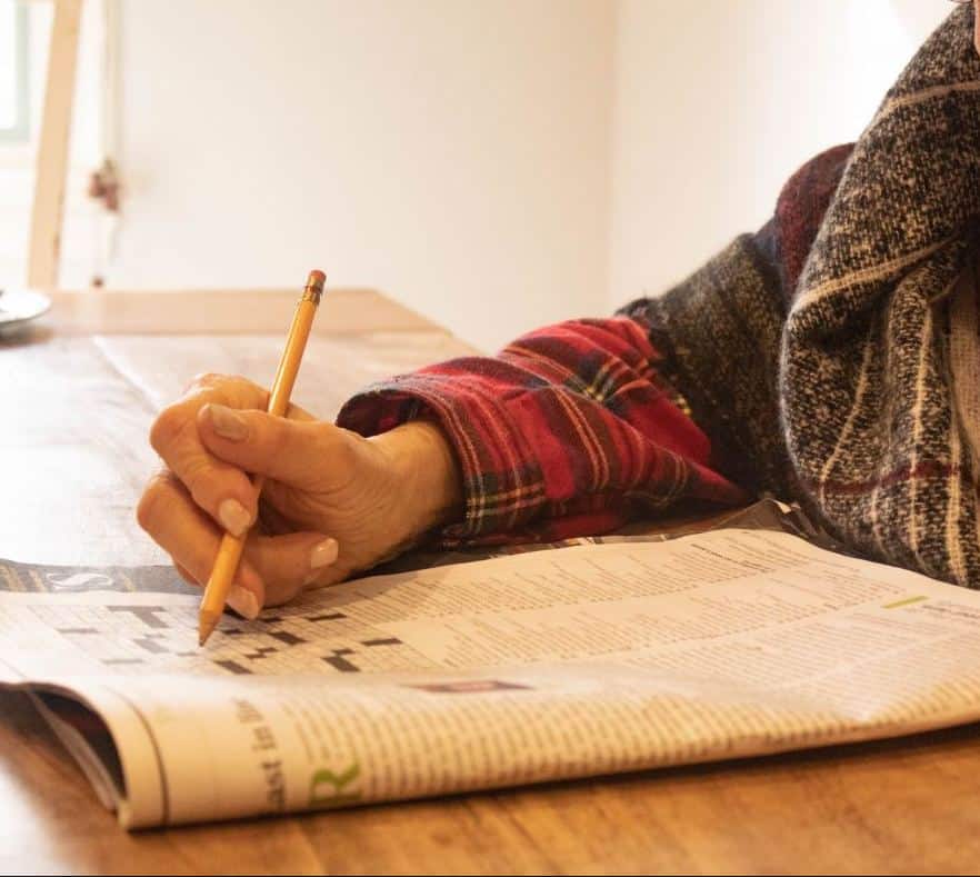 A hand is seen working on a crossword puzzle